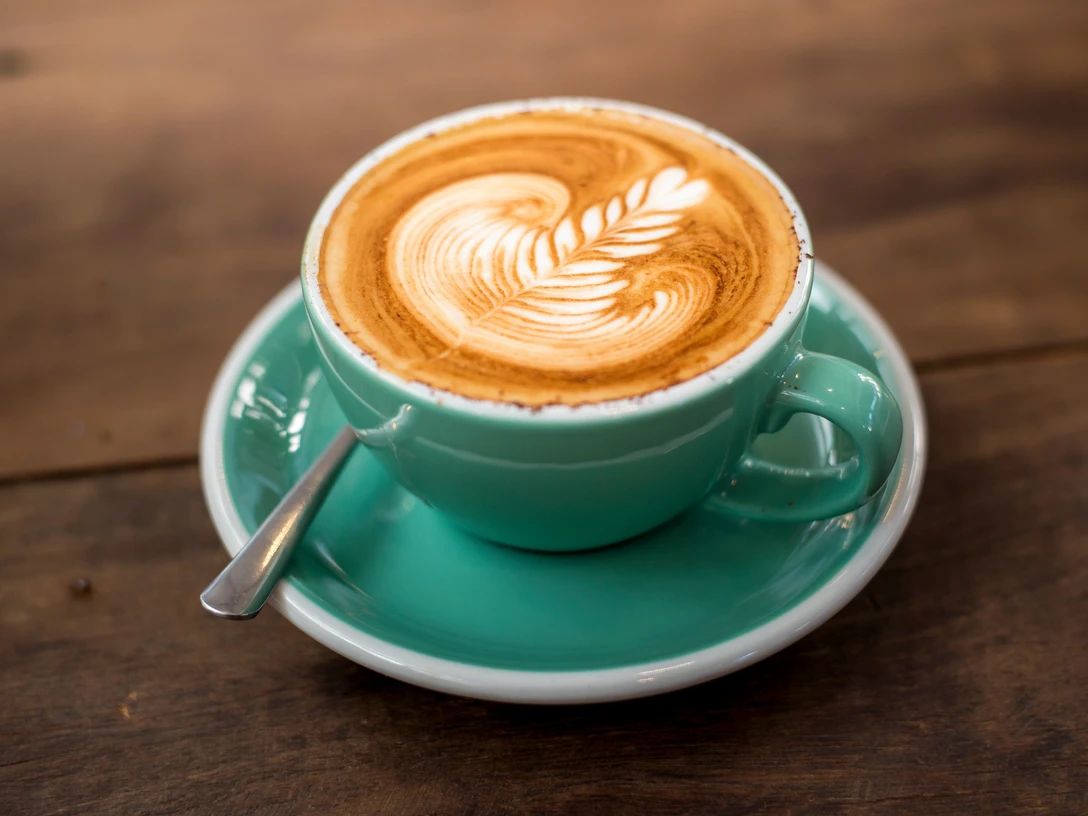 Musterbild Kaffee Eine hellgrüne Tasse mit Cappuccino, verziert mit Latte Art, steht auf einem hölzernen Tisch.