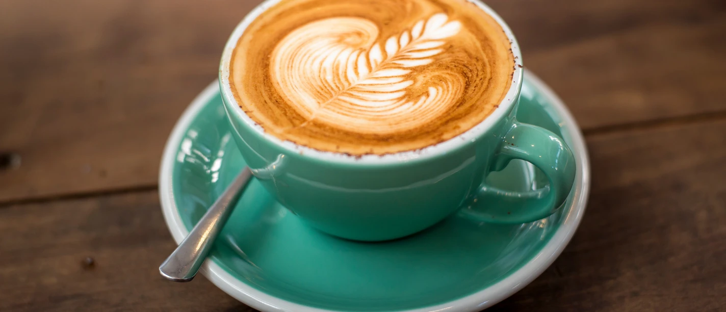 Musterbild Kaffee Eine hellgrüne Tasse mit Cappuccino, verziert mit Latte Art, steht auf einem hölzernen Tisch.