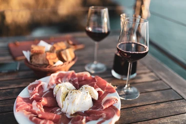 Musterbild Gastronomie Antipasti-Teller mit Prosciutto und Burrata, zwei Weingläser und Brotkorb auf Holztisch im Freien.