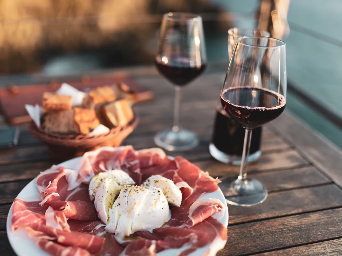 Musterbild Gastronomie Antipasti-Teller mit Prosciutto und Burrata, zwei Weingläser und Brotkorb auf Holztisch im Freien.