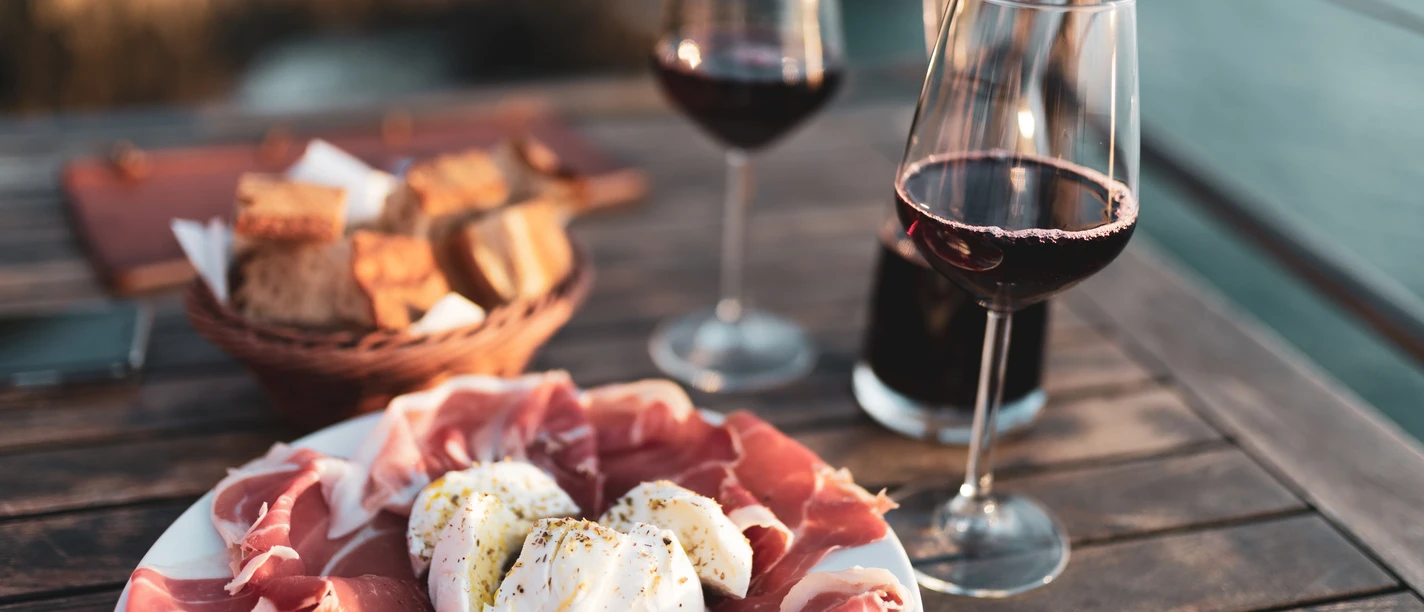 Musterbild Gastronomie Antipasti-Teller mit Prosciutto und Burrata, zwei Weingläser und Brotkorb auf Holztisch im Freien.