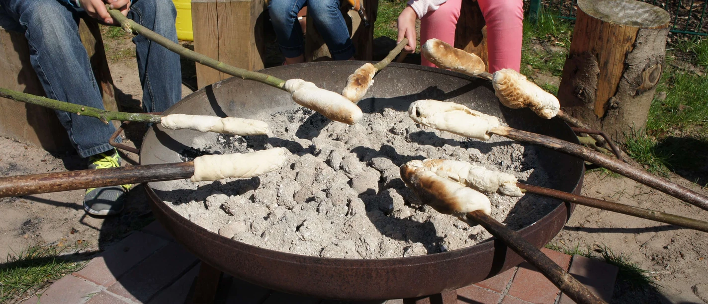 Stockbrot auf dem Abenteuerspielplatz Monheim Stockbrot backt über glühender Kohle in einer Feuerschale, umgeben von Kindern mit langen Stöcken.