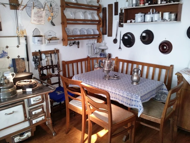 Alte Küche Gemütliche, historische Küche mit Holzstuhlgruppe, altem Herd und nostalgischem Geschirr auf Regalen.