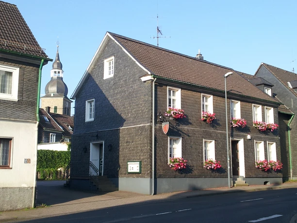Heimatmuseum Radevormwald Fachwerkhaus in sonniger Straßenszene mit blühenden Blumenkästen und Kirchturm im Hintergrund.