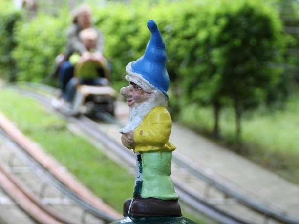 Ein bunter Gartenzwerg mit blauem Hut im Vordergrund, dahinter unscharf eine Sommerrodelbahn mit Fahrgästen.