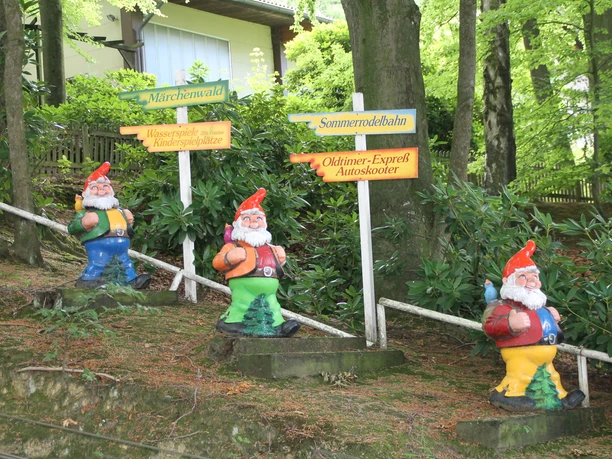 Drei bunte Zwergfiguren stehen vor Wegweisern im Grünen, die zu verschiedenen Parkattraktionen führen.