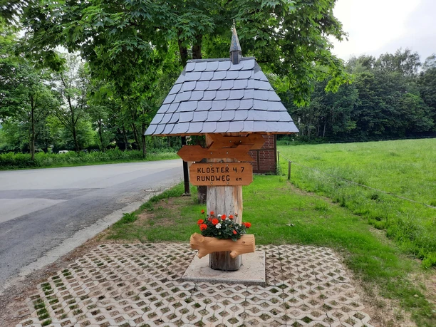 Klosterrundweg Holzwegweiser mit Schindeldach weist auf Klosterrundweg hin, umgeben von grüner Landschaft und Straße.