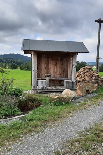 Klosterhütte Falkenhagen Eine rustikale Holzhütte mit Sitzbank am Wegesrand, umgeben von grünen Feldern und Hügeln.