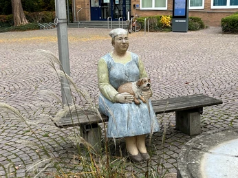 Lechner Figur Rathaus Petershagen Skulptur einer sitzenden Frau in traditioneller Kleidung mit einem Hund auf dem Schoß vor einem Gebäude.