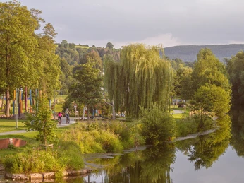 Grüne Parklandschaft mit Fluss, Bäumen und einem Kiesweg, auf dem Spaziergänger unterwegs sind.