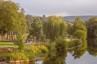 Blick auf den Emmerauenapark Grüne Parklandschaft mit Fluss, Bäumen und einem Kiesweg, auf dem Spaziergänger unterwegs sind.