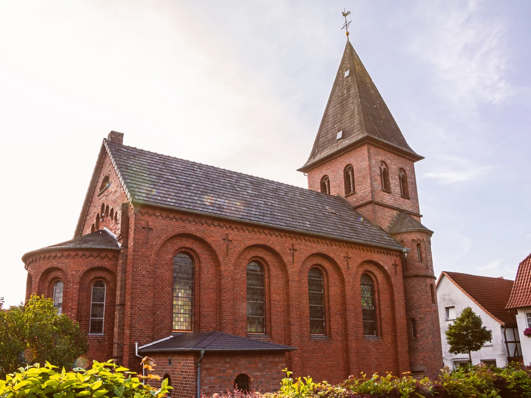 Backsteinkirche St. Johannis mit spitzem Turmdach und Rundbogenfenstern, umgeben von grünen Büschen.