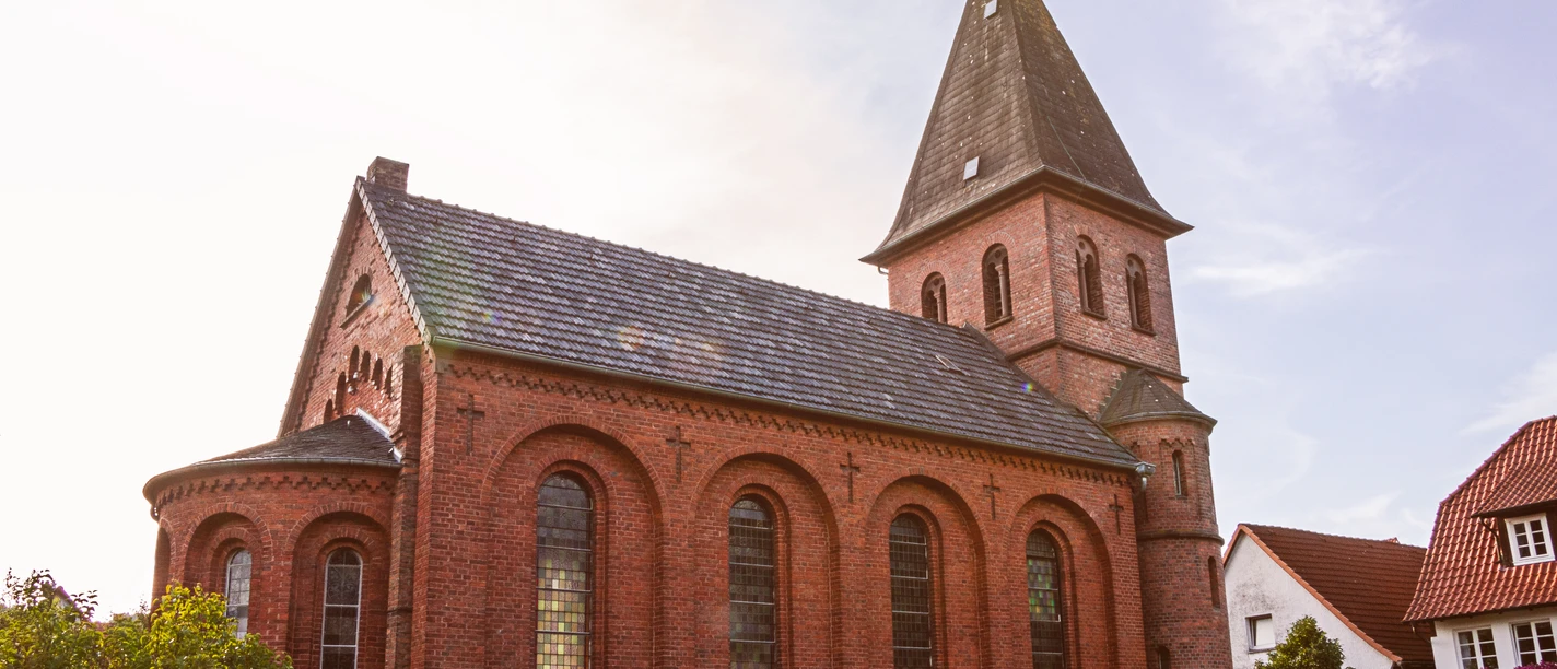 Backsteinkirche St. Johannis mit spitzem Turmdach und Rundbogenfenstern, umgeben von grünen Büschen.