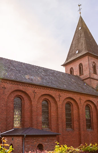 St.Johannis Kirche Backsteinkirche St. Johannis mit spitzem Turmdach und Rundbogenfenstern, umgeben von grünen Büschen.