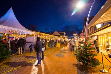 Eine Fußgängerzone bei Nacht. Auf der rechten Seite sind Geschäfte und ein Weihnachtsbaum. Auf der linken Seite befinden sich weihnachtlich geschmückte Buden.