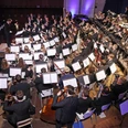 Bläserphilharmonie Aachen Bläserphilharmonie Aachen, bestehend aus vielen Musiker*innen in eleganter Abendgarderobe, die konzentriert ihre Instrumente spielen. Die Mitglieder des Orchesters sitzen in Formationen, die typische Orchesterinstrumente wie Trompeten, Hörner und Klarinetten umfassen. Der Dirigent steht vorne.