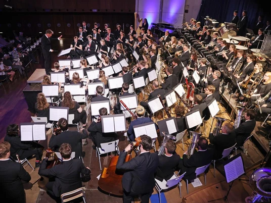 Bläserphilharmonie Aachen Bläserphilharmonie Aachen, bestehend aus vielen Musiker*innen in eleganter Abendgarderobe, die konzentriert ihre Instrumente spielen. Die Mitglieder des Orchesters sitzen in Formationen, die typische Orchesterinstrumente wie Trompeten, Hörner und Klarinetten umfassen. Der Dirigent steht vorne.