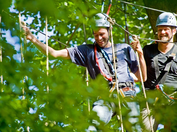 Zwei Männer mit Helmen genießen ein Kletterabenteuer im Wald, umgeben von grünen Blättern.
