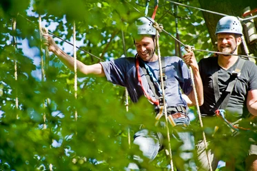 Kletterpark Interakteam_Teutoburger Wald_CCBYSA.jpg Zwei Männer mit Helmen genießen ein Kletterabenteuer im Wald, umgeben von grünen Blättern.
