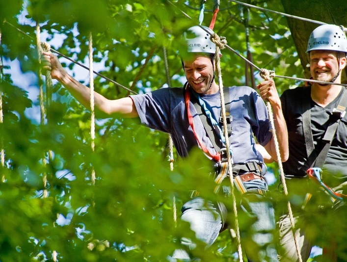 Kletterpark Interakteam_Teutoburger Wald_CCBYSA.jpg Zwei Männer mit Helmen genießen ein Kletterabenteuer im Wald, umgeben von grünen Blättern.