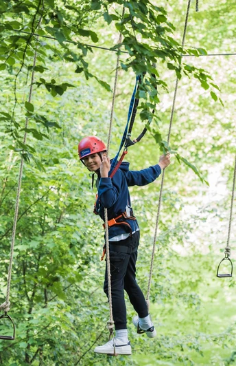 Kletterpark Interakteam_Teutoburger Wald_Leon Neufeld_CCBYSA.jpg Ein junger Kletterer balanciert auf einem Seilparcours in einem grün bewachsenen Kletterpark.
