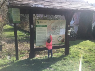 Infotafel eines Naturerlebnispfades an einer hölzernen Struktur, umgeben von grüner Landschaft. Ein Kind betrachtet die Inhalte interessiert.
