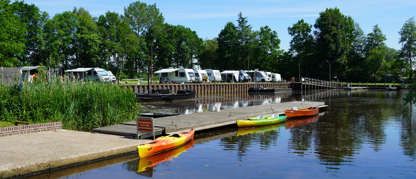 Several colorful canoes are moored on the wooden jetty of the Hammehafen, with motorhomes and trees behind them.