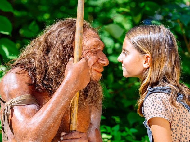 Gesichtervergleich mit MR N und Mädchen Ein Mädchen steht einem Neandertaler-Modell gegenüber, beide blicken sich intensiv in die Augen.