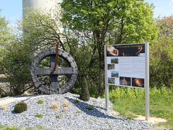 Skulptur eines Rads mit Flammenmotiv auf Steinsockel, Infotafel daneben, Bäume im Hintergrund.