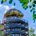 Höhlenblick-Turm mit Kalotte Aussichtsturm aus Metall mit Pilz-ähnlicher Kuppel, umgeben von Bäumen und klarem, blauem Himmel.