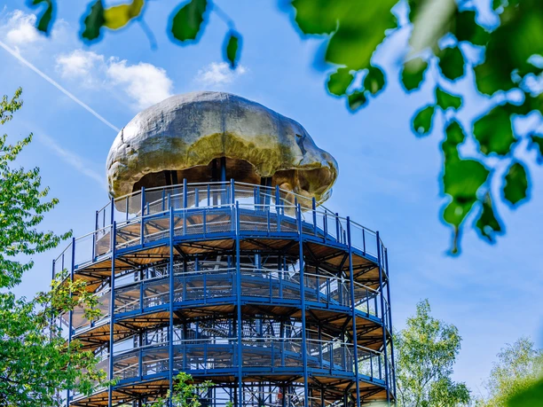 Höhlenblick-Turm mit Kalotte Aussichtsturm aus Metall mit Pilz-ähnlicher Kuppel, umgeben von Bäumen und klarem, blauem Himmel.