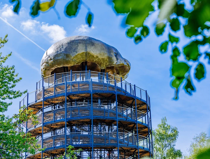 Höhlenblick-Turm mit Kalotte Aussichtsturm aus Metall mit Pilz-ähnlicher Kuppel, umgeben von Bäumen und klarem, blauem Himmel.