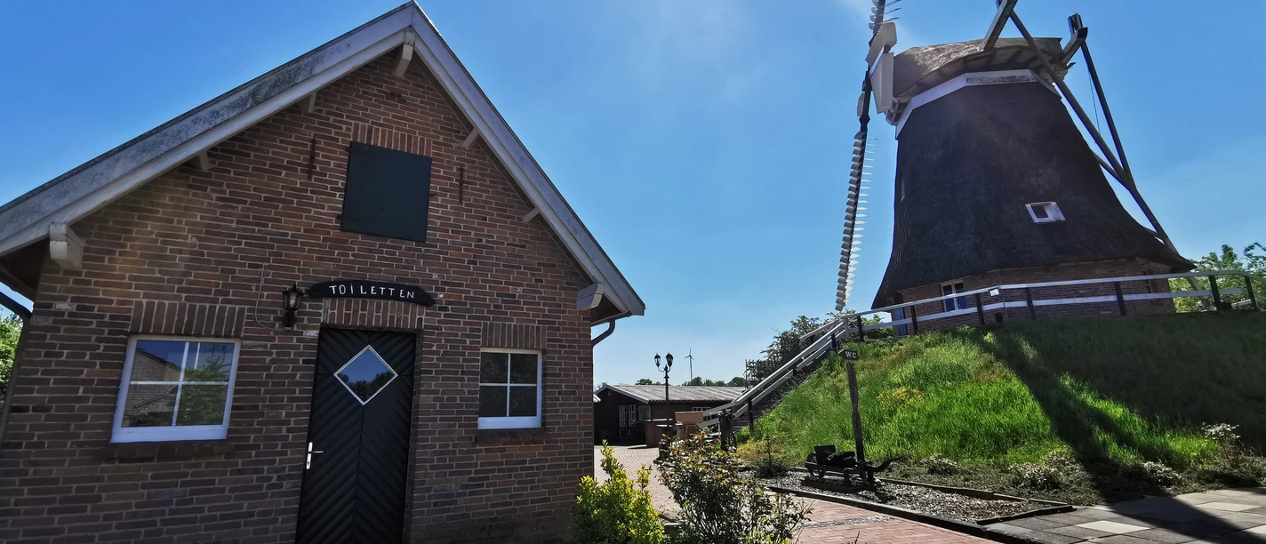 Mühle_Burlage_Rhauderfehn_31.05.21_mk (21).jpg Backsteingebäude mit schwarzer Tür und zwei Fenstern neben einer großen Windmühle mit Reetdach unter strahlendem Himmel