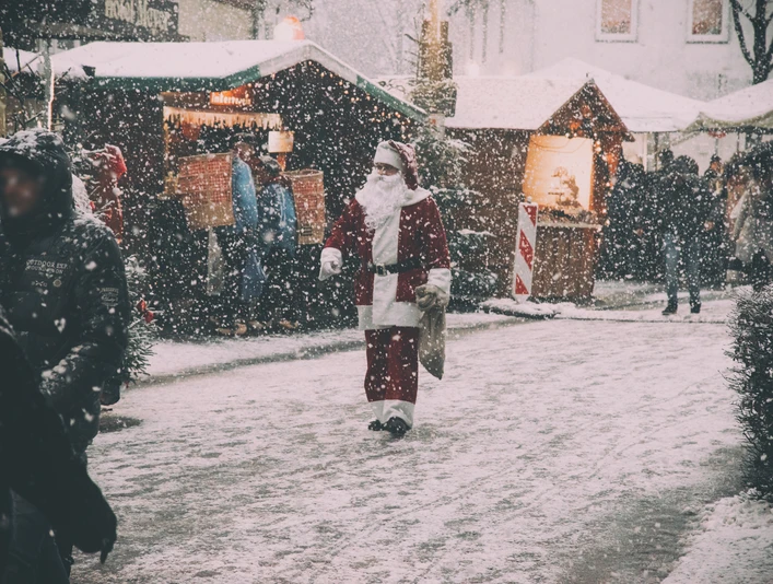 Der Weihnachtsmann geht durch einen verschneiten Weihnachtsmarkt, umgeben von Holzhütten und Besuchern.