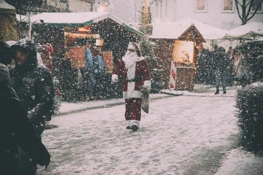 Der Weihnachtsmann geht durch einen verschneiten Weihnachtsmarkt, umgeben von Holzhütten und Besuchern.