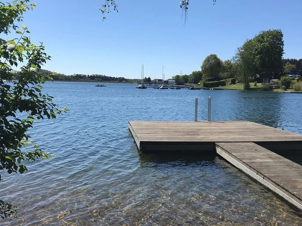Badestelle Holzsteg am klaren See mit ruhigem Wasser und Boote in der Ferne, umgeben von grüner Natur.