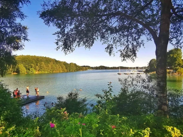 Bever-Talsperre Ein idyllischer See umgeben von Bäumen, mit Schwimmern auf einem Badesteg bei sonnigem Wetter.