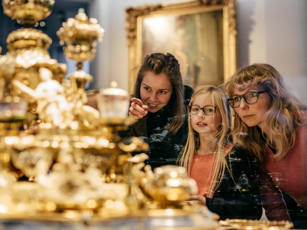 SKD_Schloss_Image_MG_4375_davidpinzer_kleiner_1611.jpg Besucher betrachten goldene Kunstwerke im Museum, reflektiert in einer Glasvitrine.