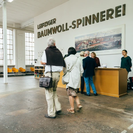 W hali oznaczonej jako "Leipziger Baumwoll-Spinnerei" niektórzy odwiedzający stoją przy stoliku informacyjnym, w tle widać duże okna i żółty rząd siedzeń, kulturę przemysłową, sztukę.