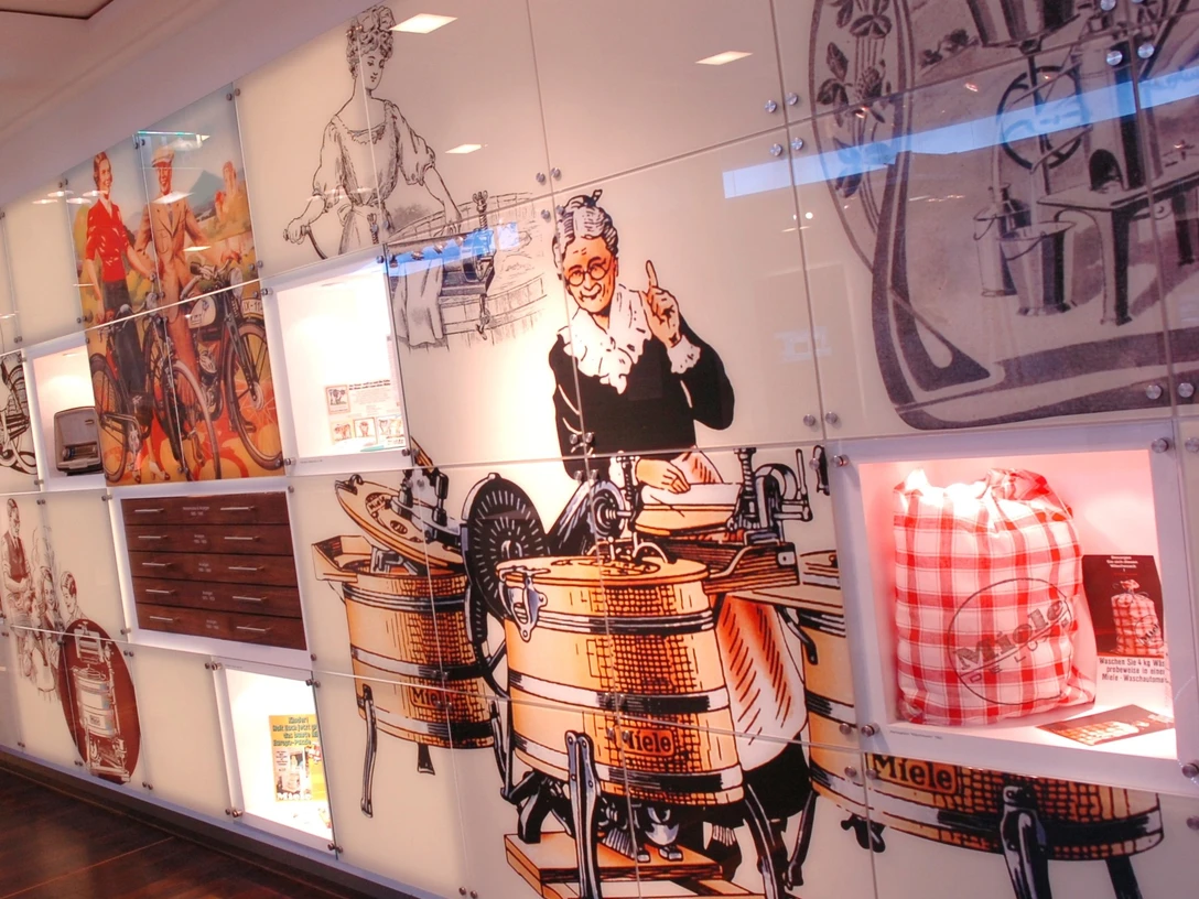 2006-206_06.jpg Retro-Illustrationen alter Miele-Waschmaschinen verzieren eine moderne weiße Wand in einem Ausstellungsraum.