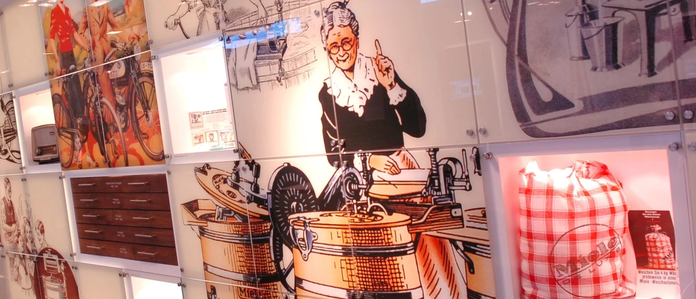 2006-206_06.jpg Retro-Illustrationen alter Miele-Waschmaschinen verzieren eine moderne weiße Wand in einem Ausstellungsraum.