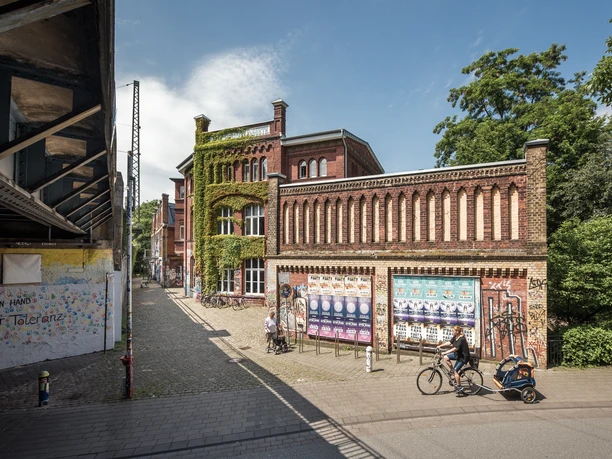 Weberei Gütersloh Roter Backsteinbau mit grauer Fassade, bewachsene Wand, Fahrradfahrende Personen, blauer Himmel.