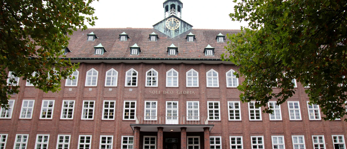 Backsteingebäude mit Uhrturm und Schriftzug "Soli Deo Gloria", umgeben von Bäumen und Autos im Vordergrund.