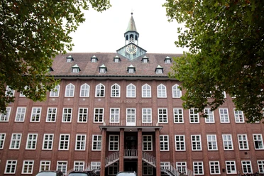 ESG Gütersloh Backsteingebäude mit Uhrturm und Schriftzug "Soli Deo Gloria", umgeben von Bäumen und Autos im Vordergrund.
