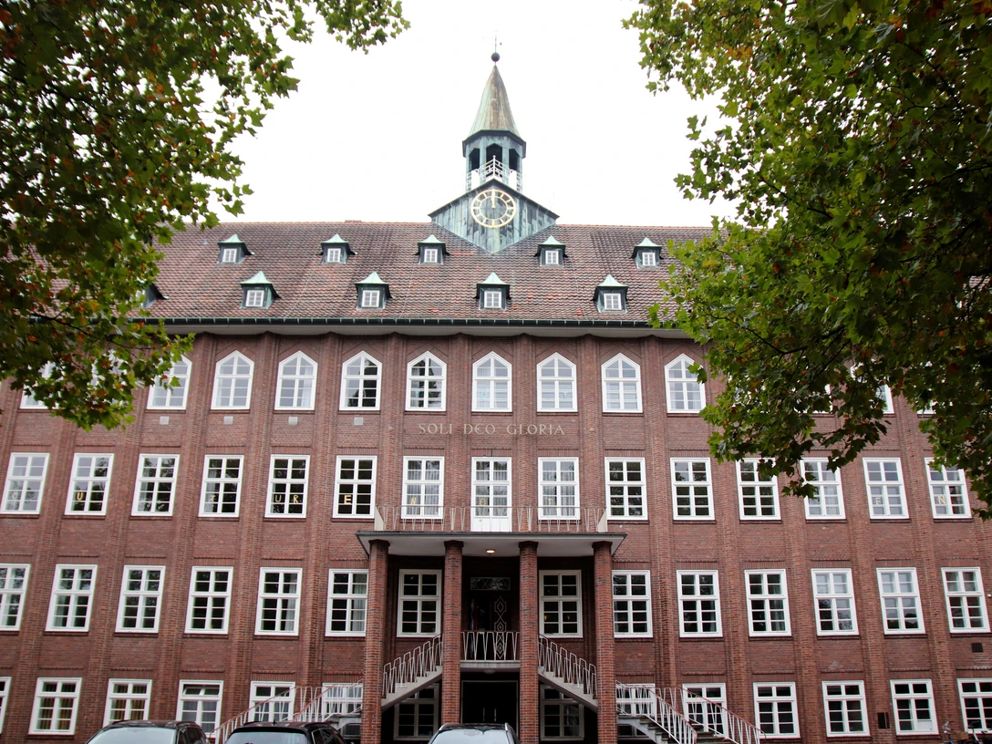 ESG Gütersloh Backsteingebäude mit Uhrturm und Schriftzug "Soli Deo Gloria", umgeben von Bäumen und Autos im Vordergrund.