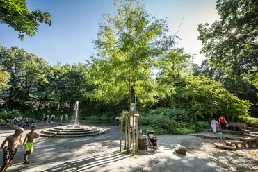 Mohnspark Kinder spielen an einem Wasserspielplatz mit Springbrunnen, umgeben von grünen Bäumen im Sonnenschein.