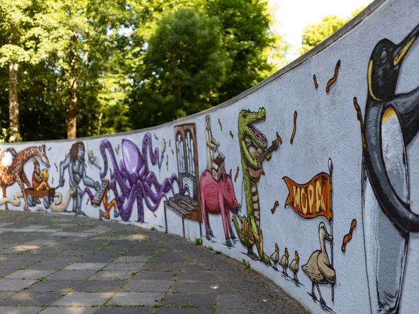 Bunte Tier-Graffiti schmücken eine geschwungene Mauer im Freien, darunter ein Pinguin und ein Dinosaurier.
