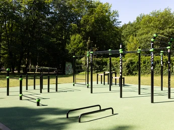 Mohns Park Gütersloh Barrierefreier Fitnesspark mit Outdoor-Trainingsgeräten, umgeben von Bäumen und Rasenflächen im Sonnenschein.