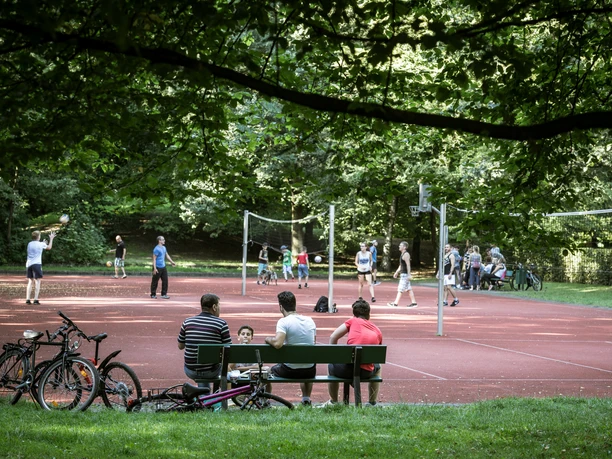 Personen spielen Volleyball auf einem Sportplatz im Park, Fahrräder und Zuschauer im Vordergrund.