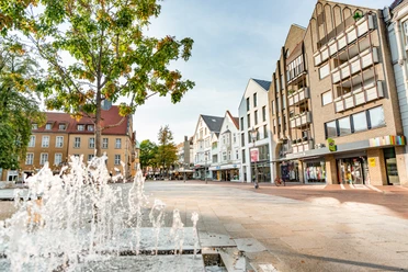 Belebter Platz mit Wasserspielen, umgeben von modernen Gebäuden und Bäumen unter blauem Himmel.